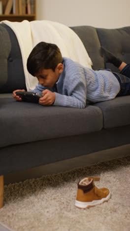 Vertical-Video-Of-Young-Boy-Lying-On-Sofa-At-Home-Playing-Game-Or-Streaming-Onto-Handheld-Gaming-Device